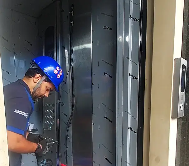 Técnico da Thopp Elevadores, de capacete azul, instalando a botoeira em uma cabine nova de elevador ainda com película protetora no inox