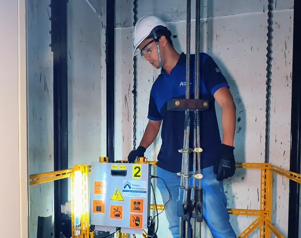 Técnico da Thopp Elevadores, de capacete, óculos e luvas de proteção, fazendo a manutenção no topo da cabine, junto à botoeira de inspeção e ao guarda-corpo amarelo