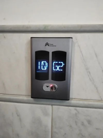 Indicador de pavimento duplo com displays digitais e botão de chamada, instalado em parede de mármore