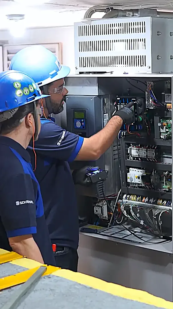 Dois técnicos da Thopp Elevadores, de capacete azul, conferindo o quadro de comando de um elevador na casa de máquinas