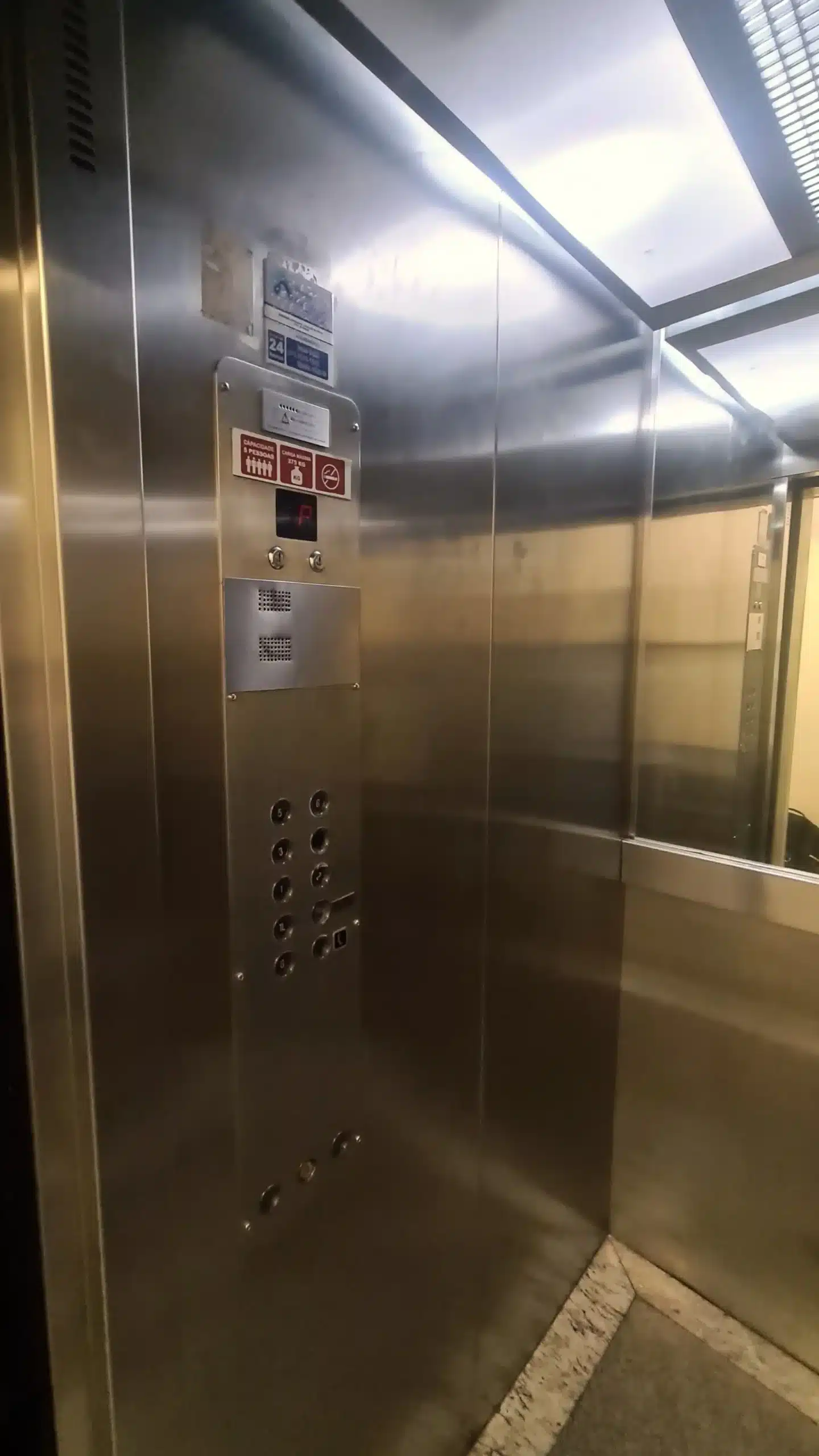 Interior da cabine em inox antes da renovação, com a botoeira anterior, indicador de andar de segmentos vermelhos e placas de aviso coladas