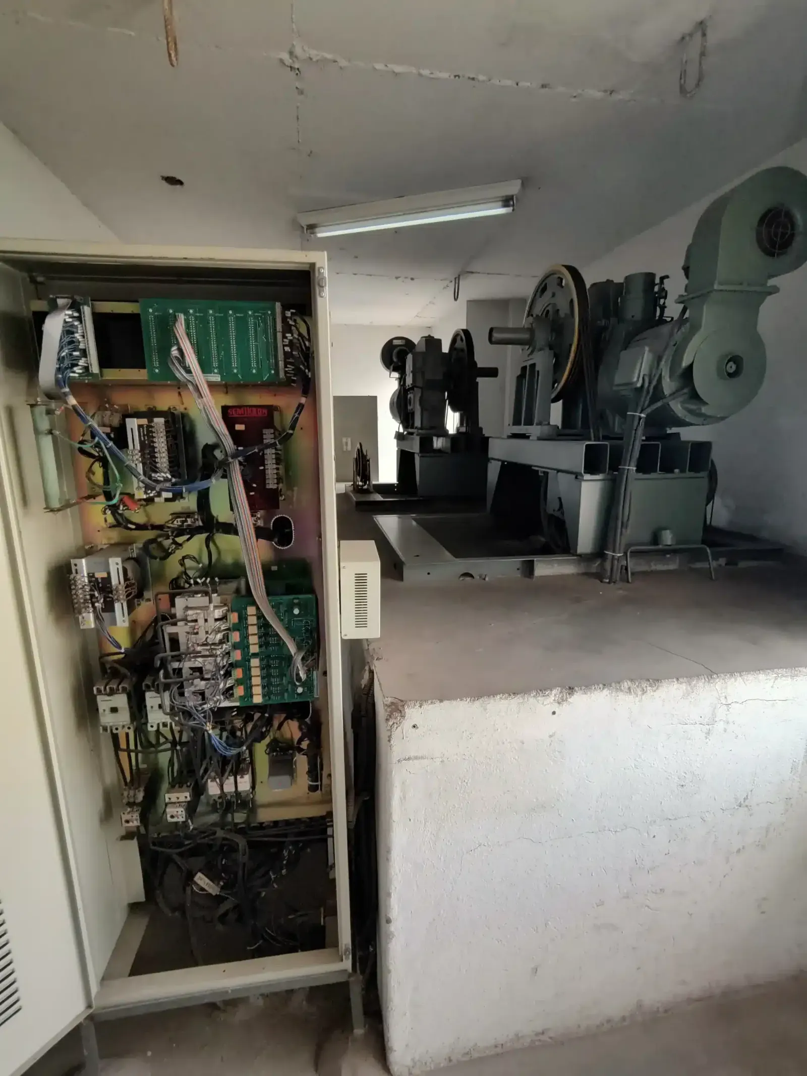 Casa de máquinas do edifício na Rua Manaus antes da modernização, com o quadro de comando anterior aberto, placas eletrônicas e fiação expostas, ao lado das máquinas de tração
