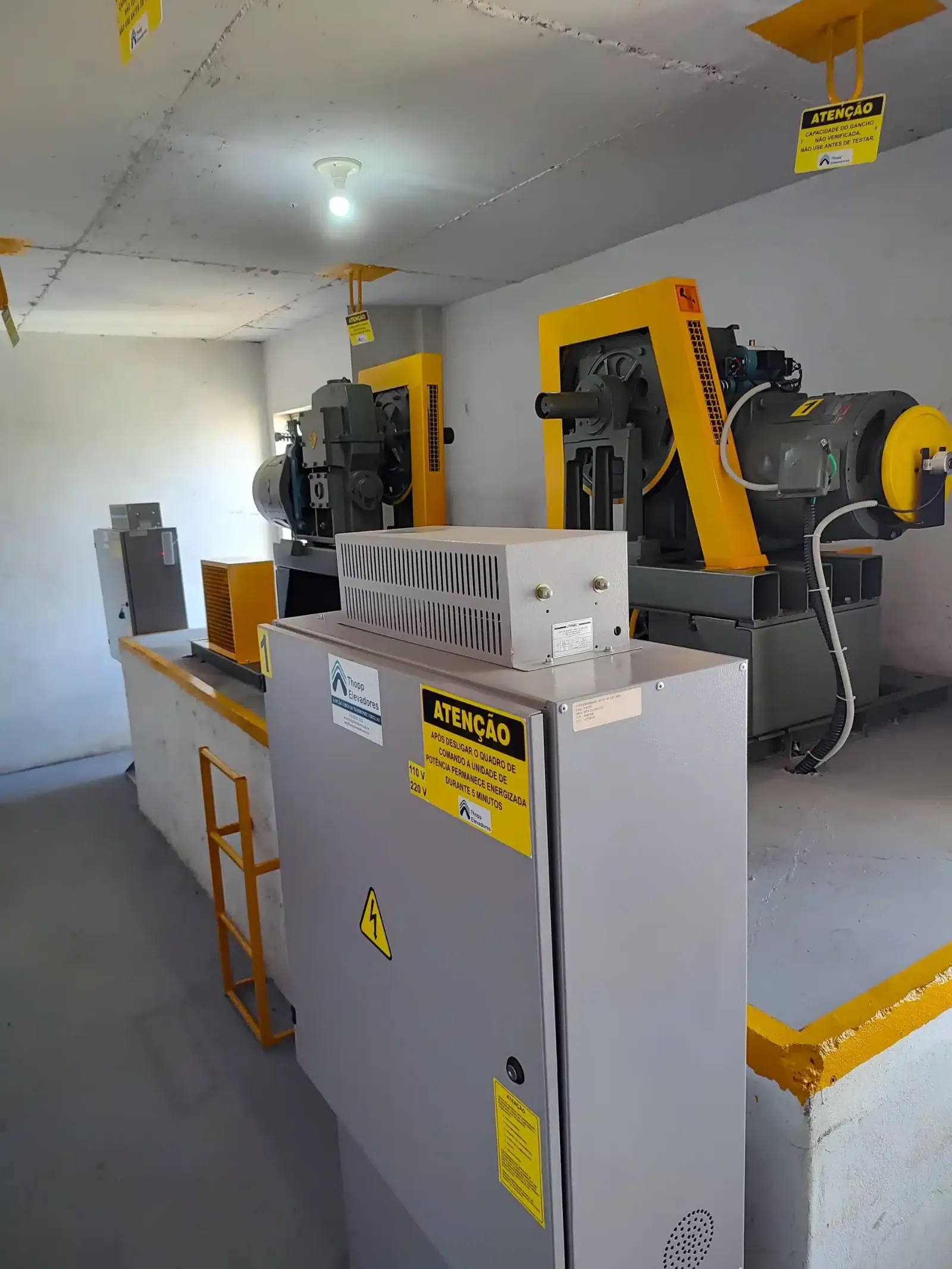 A mesma casa de máquinas depois da modernização, com quadro de comando novo, fechado e sinalizado, máquinas de tração com proteções amarelas, escada de acesso e bordas pintadas de amarelo