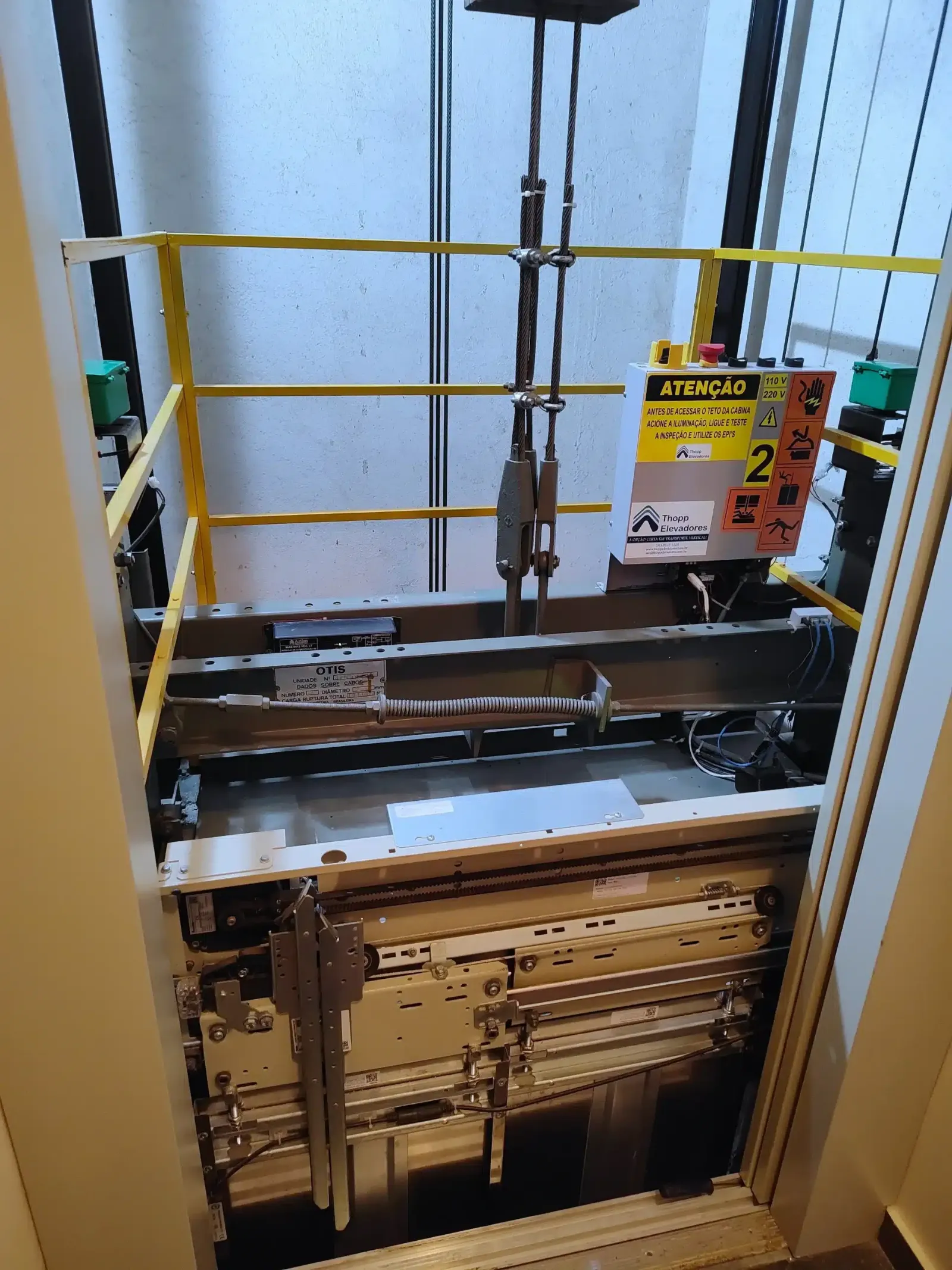 Topo da cabine do mesmo elevador depois da modernização, com guarda-corpo amarelo, caixa de inspeção sinalizada da Thopp e operador de porta novo; a placa de dados da armação continua no lugar